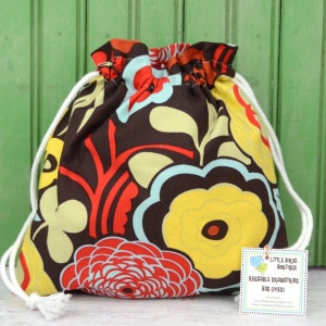 May include: A brown drawstring bag with a floral pattern in shades of red, yellow, blue, and green. The bag is made of fabric and has a white drawstring. The bag has a tag attached to it that reads "Little Birds Boutique Reusable Drawstring Bag (Med)".