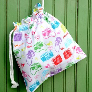 May include: White drawstring bag with a colorful camera print pattern. The bag has a white drawstring and the text "I ♥  pix" and "click" is repeated on the bag.