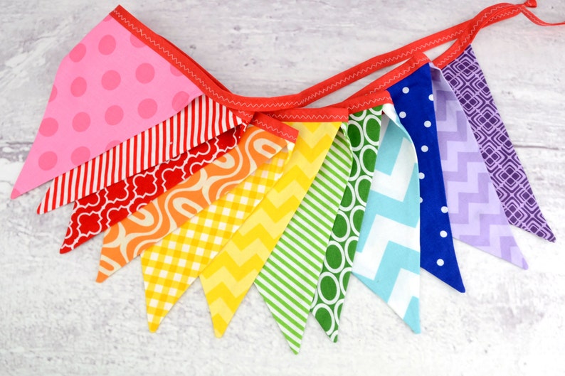 May include: A rainbow fabric banner with 10 triangular flags. The flags are in various colours and patterns, including pink with white polka dots, red with white stripes, orange with a wavy pattern, yellow with a checkered pattern, green with white stripes, blue with white polka dots, and purple with a geometric pattern.