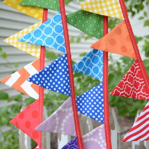 May include: A colourful fabric bunting with triangular flags in various patterns, including polka dots, gingham, and geometric designs. The flags are strung together with red ribbon.