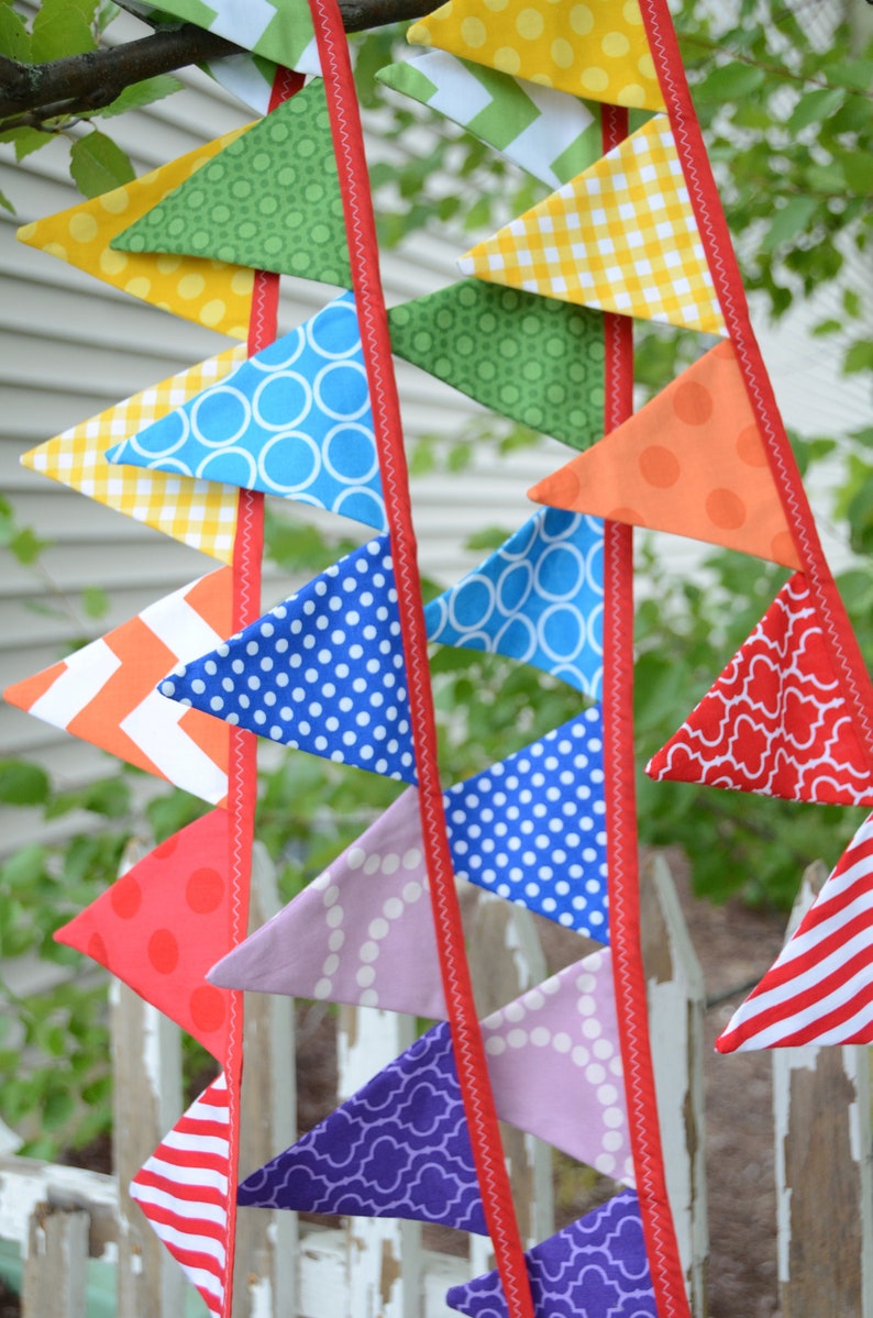 May include: A colourful fabric bunting with triangular flags in various patterns, including polka dots, gingham, and geometric designs. The flags are strung together with red ribbon.