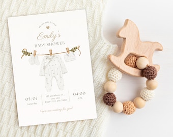 Puede incluir: Una invitación a un baby shower con el texto "Emily's Baby Shower" y una ilustración en acuarela de ropa de bebé colgada en un tendedero. Un sonajero para bebé con forma de caballo balancín de madera con cuentas de ganchillo y madera está junto a la invitación.
