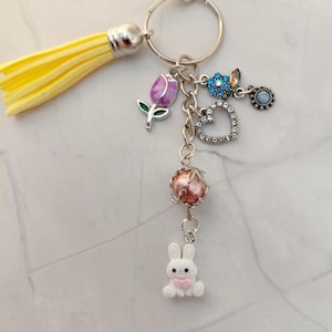 May include: A silver keychain with a yellow tassel and various charms. Charms include a purple tulip, a rhinestone heart, a blue flower, a pink pearl, a small blue charm, and a white bunny with a pink bow.