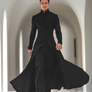 以下が含まれることがあります： ハイカラーと流れるようなデザインの黒いロングコート。コートは黒いパンツの上に着用されています。モデルは明るい廊下を歩いています。コートのデザインは、クラシックなスタイルのモダンな解釈です。