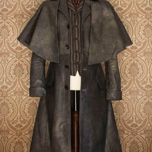 May include: A dark gray, vintage-style coat with a cape, displayed on a wooden mannequin. The coat features a long design with buttons and pockets. Underneath, a striped vest and patterned scarf are visible.