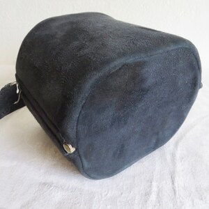 Puede incluir: Un bolso cil&iacute;ndrico de ante negro con un acabado mate texturizado. El bolso presenta un panel lateral con un peque&ntilde;o bot&oacute;n a presi&oacute;n met&aacute;lico de tono plateado y una correa de ante negro a juego sujeta a una anilla de metal plateado.
