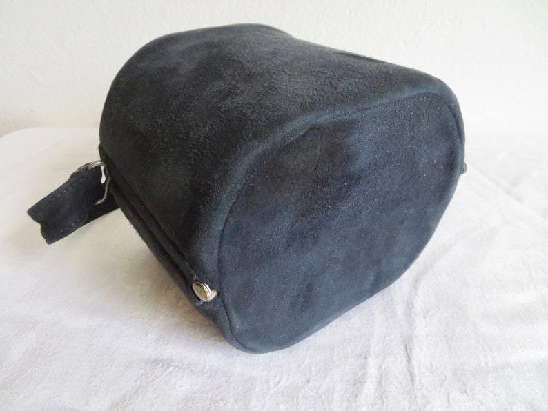 Puede incluir: Un bolso cil&iacute;ndrico de ante negro con un acabado mate texturizado. El bolso presenta un panel lateral con un peque&ntilde;o bot&oacute;n a presi&oacute;n met&aacute;lico de tono plateado y una correa de ante negro a juego sujeta a una anilla de metal plateado.
