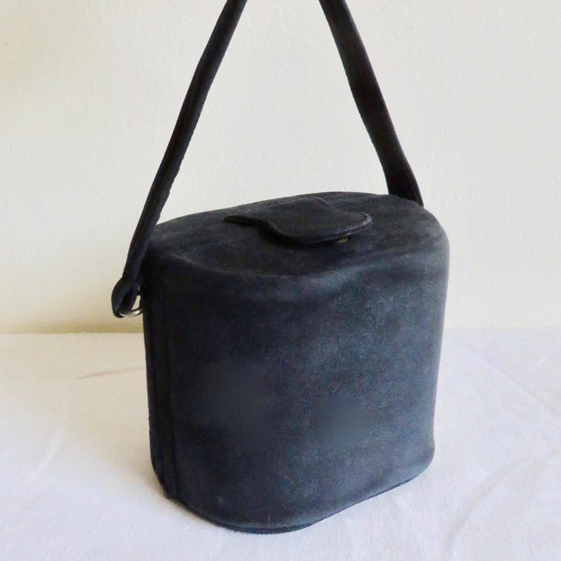 Puede incluir: Un bolso de mano negro estilo cubo con una textura suave y mate. El bolso presenta un cuerpo cil&iacute;ndrico redondeado, un asa superior &uacute;nica sujeta a los lados con anillos de metal y un peque&ntilde;o cierre de solapa redondeado centrado en la parte superior.