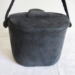 Puede incluir: Un bolso de mano negro estilo cubo con un acabado texturizado similar al ante. El bolso presenta una base estructurada y redondeada, un cierre de solapa superior con una leng&uuml;eta central y una sola correa negra sujeta con anillas met&aacute;licas.