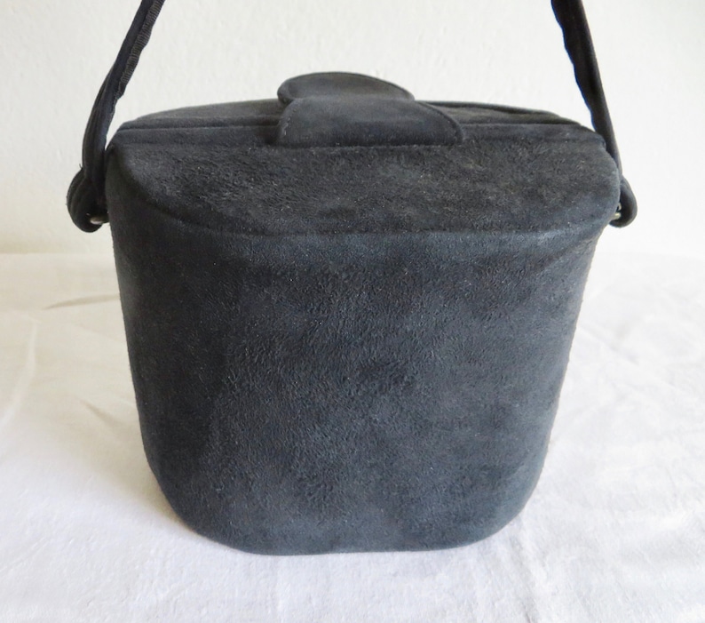 Puede incluir: Un bolso de mano negro estilo cubo con un acabado texturizado similar al ante. El bolso presenta una base estructurada y redondeada, un cierre de solapa superior con una leng&uuml;eta central y una sola correa negra sujeta con anillas met&aacute;licas.