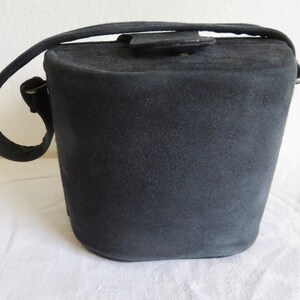 Puede incluir: Un bolso de mano negro estilo cubo con un acabado mate y texturizado. El bolso presenta una base redondeada y estructurada, un asa superior &uacute;nica y un cierre de solapa asegurado con un peque&ntilde;o bot&oacute;n met&aacute;lico de aspecto suave y aterciopelado.