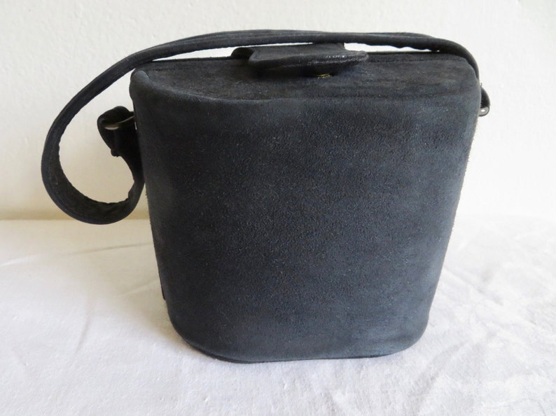 Puede incluir: Un bolso de mano negro estilo cubo con un acabado mate y texturizado. El bolso presenta una base redondeada y estructurada, un asa superior &uacute;nica y un cierre de solapa asegurado con un peque&ntilde;o bot&oacute;n met&aacute;lico de aspecto suave y aterciopelado.