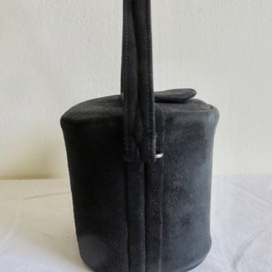 Puede incluir: Un bolso cil&iacute;ndrico de ante negro con un asa vertical &uacute;nica unida a los lados. El bolso tiene una base circular plana y un cierre de tapa superior. El exterior est&aacute; hecho de un material negro mate con costuras visibles a lo largo del asa.