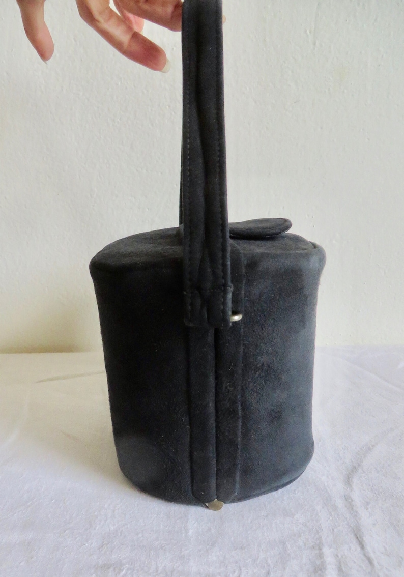 Puede incluir: Un bolso cil&iacute;ndrico de ante negro con un asa vertical &uacute;nica unida a los lados. El bolso tiene una base circular plana y un cierre de tapa superior. El exterior est&aacute; hecho de un material negro mate con costuras visibles a lo largo del asa.