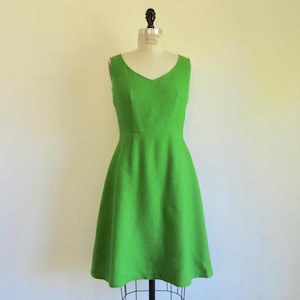 Puede incluir: Un vestido verde brillante, sin mangas, con escote en V y falda acampanada. El vestido se exhibe en un maniquí. La tela tiene una textura sutil y el estilo está inspirado en lo vintage.