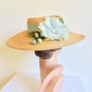 Peut inclure: Un chapeau de paille &agrave; larges bords avec une fleur bleu clair et des feuilles vertes. Le chapeau a un ruban vert clair autour de la couronne.