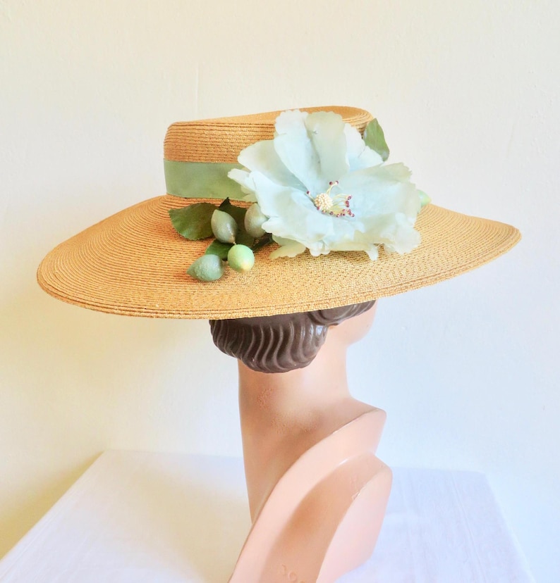 Peut inclure: Un chapeau de paille &agrave; larges bords avec une fleur bleu clair et des feuilles vertes. Le chapeau a un ruban vert clair autour de la couronne.