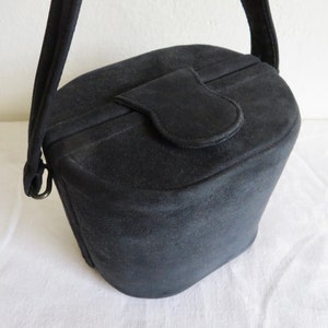 Puede incluir: Un bolso de mano negro estilo cubo con acabado mate, que presenta un cierre de solapa superior redondeado y una sola correa para el hombro unida a los lados con anillos de metal. El bolso tiene una forma cil&iacute;ndrica estructurada con base plana.