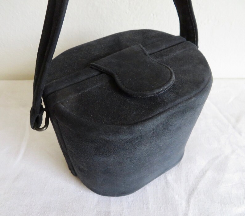 Puede incluir: Un bolso de mano negro estilo cubo con acabado mate, que presenta un cierre de solapa superior redondeado y una sola correa para el hombro unida a los lados con anillos de metal. El bolso tiene una forma cil&iacute;ndrica estructurada con base plana.