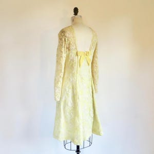 May include: A pale yellow long-sleeved dress featuring an intricate floral lace overlay. The garment has a deep scoop back neckline accented with a prominent yellow bow detail at the center, falling to a knee-length hem on a dress form.