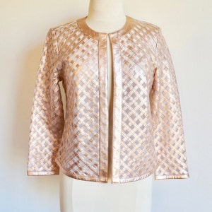 Puede incluir: Chaqueta rosa dorada con un patrón de celosía, exhibida en un maniquí. La chaqueta tiene un cuello redondo y una abertura vertical en la parte delantera. Las mangas son largas y la chaqueta parece estar hecha de un material metálico.