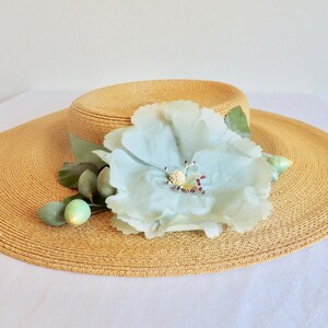 Peut inclure: Un chapeau de paille &agrave; larges bords avec une fleur bleu clair et des feuilles vertes. Le chapeau est de couleur brun clair et a une texture tiss&eacute;e.