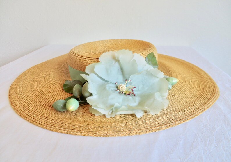 Peut inclure: Un chapeau de paille &agrave; larges bords avec une fleur bleu clair et des feuilles vertes. Le chapeau est de couleur brun clair et a une texture tiss&eacute;e.