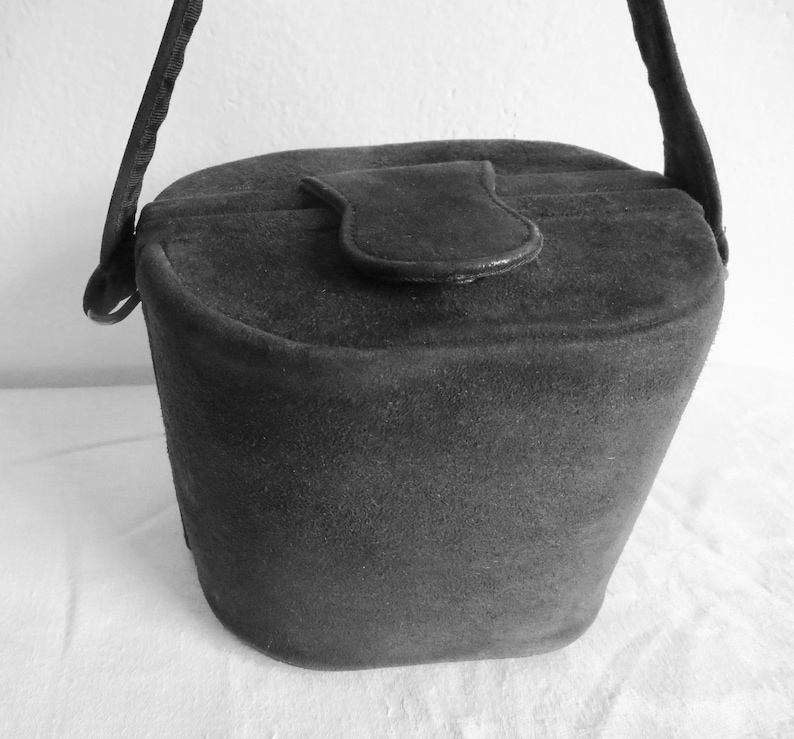 Puede incluir: Un bolso de mano negro estilo cubo con una textura suave y mate. El bolso presenta un cierre de solapa superior redondeado con una leng&uuml;eta central y una sola correa para el hombro unida a los lados. La forma es c&oacute;nica, m&aacute;s ancha arriba.