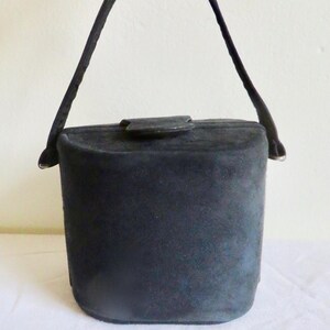 Puede incluir: Un bolso de mano tipo cubo en ante negro con una forma cil&iacute;ndrica estructurada, un asa superior &uacute;nica y un peque&ntilde;o cierre de solapa en la parte superior. El bolso presenta una textura mate, una base plana y lados redondeados.