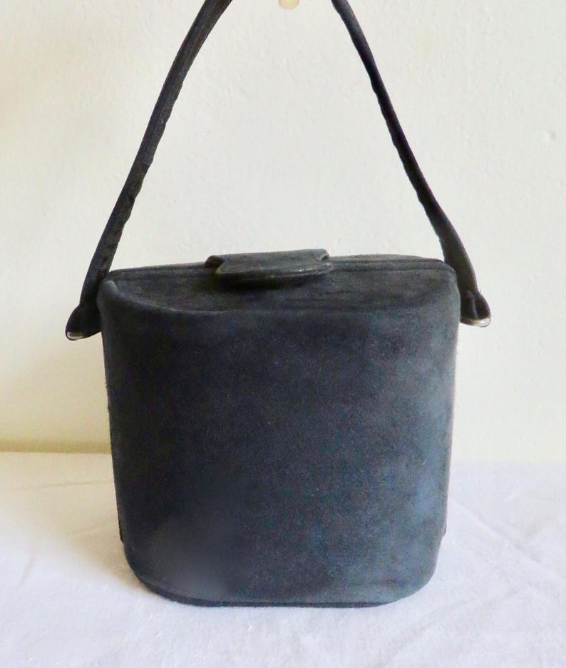 Puede incluir: Un bolso de mano tipo cubo en ante negro con una forma cil&iacute;ndrica estructurada, un asa superior &uacute;nica y un peque&ntilde;o cierre de solapa en la parte superior. El bolso presenta una textura mate, una base plana y lados redondeados.