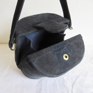 Puede incluir: Un bolso de mano de ante negro con una silueta estructurada semicircular y una sola asa superior. El bolso cuenta con un cierre de solapa asegurado por un bot&oacute;n de presi&oacute;n de metal dorado, con fuelles interiores visibles en los lados.