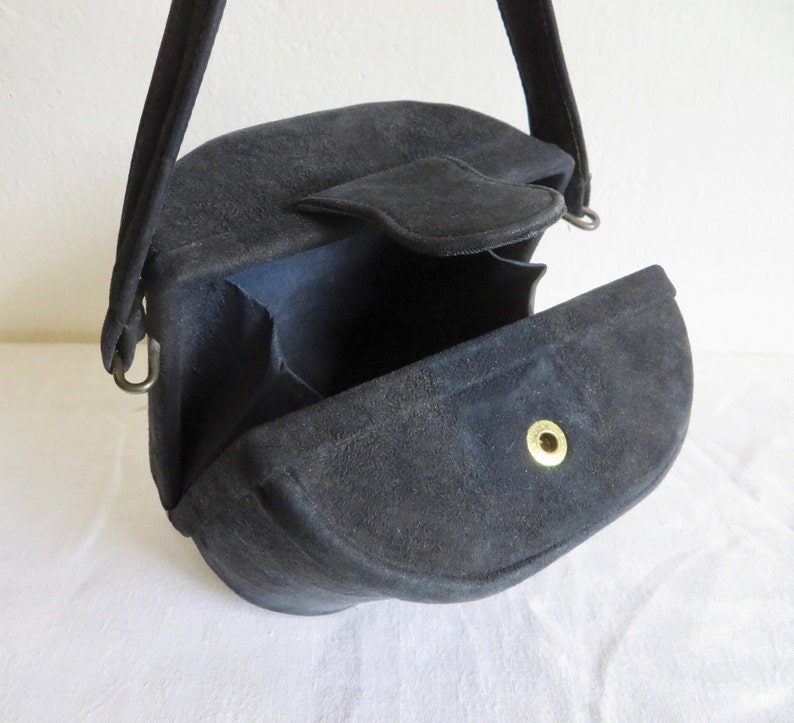 Puede incluir: Un bolso de mano de ante negro con una silueta estructurada semicircular y una sola asa superior. El bolso cuenta con un cierre de solapa asegurado por un bot&oacute;n de presi&oacute;n de metal dorado, con fuelles interiores visibles en los lados.
