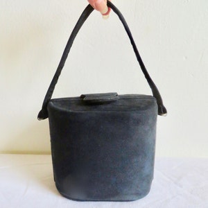 Puede incluir: Un bolso de mano negro estilo cubo con una textura suave y mate. El bolso cuenta con un asa arqueada &uacute;nica unida a los lados y un peque&ntilde;o cierre de solapa centrado en la parte superior. La forma general es cil&iacute;ndrica con una base plana y redondeada.