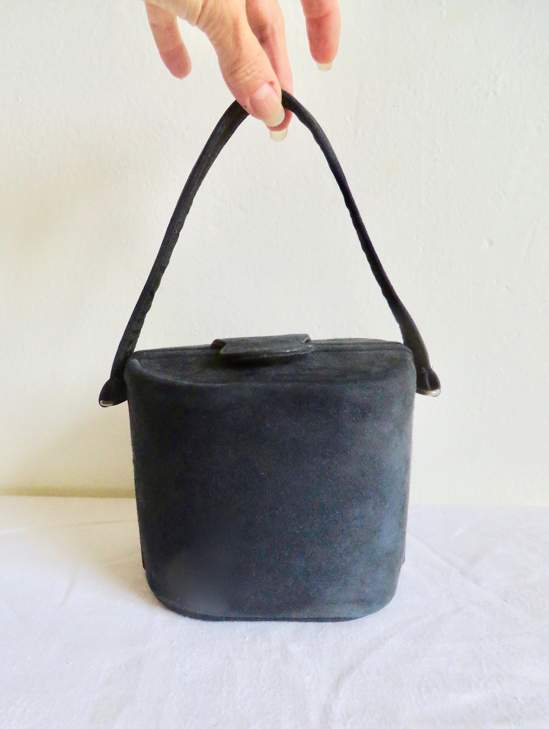 Puede incluir: Un bolso de mano negro estilo cubo con una textura suave y mate. El bolso cuenta con un asa arqueada &uacute;nica unida a los lados y un peque&ntilde;o cierre de solapa centrado en la parte superior. La forma general es cil&iacute;ndrica con una base plana y redondeada.