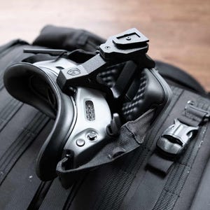 May include: A black and silver VR headset with a black mounting bracket is attached to a black backpack. The headset has a curved design with a padded area for the eyes. The backpack has multiple straps and a metal buckle.