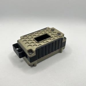 Może przedstawiać: Prostokątne, beżowe urządzenie z czarnym, teksturowanym wierzchem i bokami. Wierzch ma wzór plastra miodu i prostokątny otwór. Mały czarny element wystaje z jednej strony. Urządzenie wydaje się być wykonane z tworzywa sztucznego.