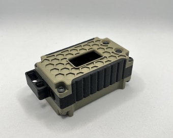 May include: A rectangular, tan-colored device with a black, textured top and sides. The top features a honeycomb pattern and a rectangular opening. A small black component extends from one side. The device appears to be made of plastic.