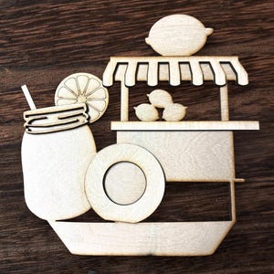 May include: Wooden laser-cut design featuring a lemonade stand, a mason jar with a straw, and a lemon slice. The stand has a lemon on top and lemons inside. The design is made of light-colored wood and is set against a dark wood background.