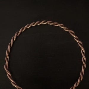 Op de afbeelding: Een koperkleurige, gedraaide metalen armband. De armband is cirkelvormig met een sluiting. Het ontwerp heeft een gevlochten of touwachtig patroon. De sieraden zijn geplaatst tegen een donkere achtergrond.