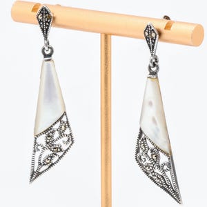 May include: Silver-tone drop earrings with a geometric design. Each earring has a pearlescent, triangular inlay and a detailed filigree pattern. The earrings are displayed on a gold-coloured jewelry stand against a white background.