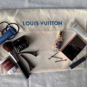 May include: An assortment of beauty and personal care products arranged on a beige Louis Vuitton pouch. Items include a makeup brush, hair clips, hair ties, perfume, mascara, and skincare products. The pouch has the brand name printed in blue.