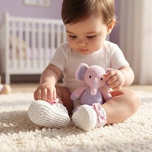 May include: A pair of white crocheted baby booties featuring a small pink bow on the front of each shoe, paired with a small, light purple and pink crocheted elephant stuffed animal with dark eyes and a trunk, held by a baby in a white bodysuit.