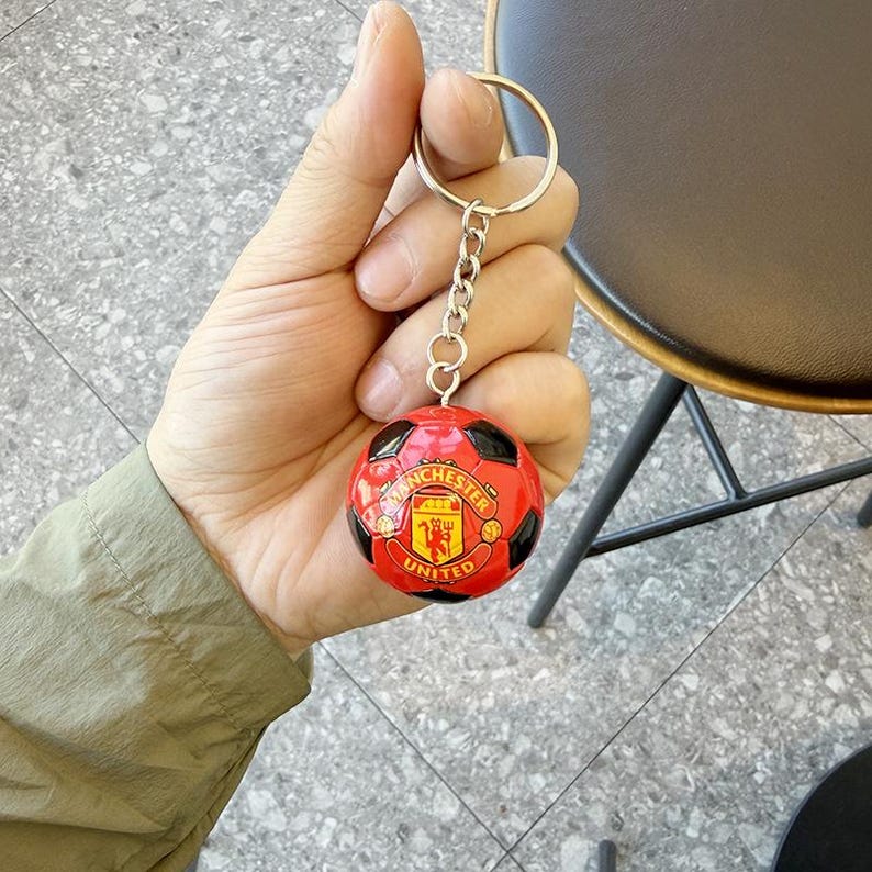 Puede incluir: Un llavero redondo con un dise&ntilde;o de bal&oacute;n de f&uacute;tbol en miniatura en rojo y negro. El centro del bal&oacute;n muestra el logotipo del club de f&uacute;tbol Manchester United en amarillo y rojo. El llavero est&aacute; unido a una anilla de metal de tono plateado.