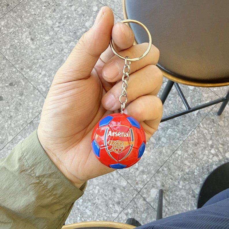 Puede incluir: Un llavero esf&eacute;rico con un dise&ntilde;o de bal&oacute;n de f&uacute;tbol rojo y azul y el logotipo del club de f&uacute;tbol Arsenal impreso en la parte frontal. El bal&oacute;n est&aacute; unido a una anilla de metal plateado mediante una cadena corta, creando un accesorio deportivo.