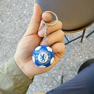 Puede incluir: Un llavero esf&eacute;rico con un dise&ntilde;o de bal&oacute;n de f&uacute;tbol en miniatura con paneles azules y blancos. El centro del bal&oacute;n muestra el logotipo del Chelsea Football Club, que incluye un le&oacute;n azul sobre fondo blanco con el texto Chelsea Football Club.