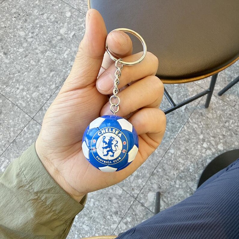 Puede incluir: Un llavero esf&eacute;rico con un dise&ntilde;o de bal&oacute;n de f&uacute;tbol en miniatura con paneles azules y blancos. El centro del bal&oacute;n muestra el logotipo del Chelsea Football Club, que incluye un le&oacute;n azul sobre fondo blanco con el texto Chelsea Football Club.