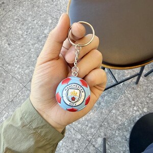Puede incluir: Un llavero esf&eacute;rico con un dise&ntilde;o de bal&oacute;n de f&uacute;tbol en azul claro y rojo, con el logotipo del club de f&uacute;tbol Manchester City impreso en la parte frontal. El bal&oacute;n est&aacute; unido a una anilla de metal plateado mediante una cadena corta.
