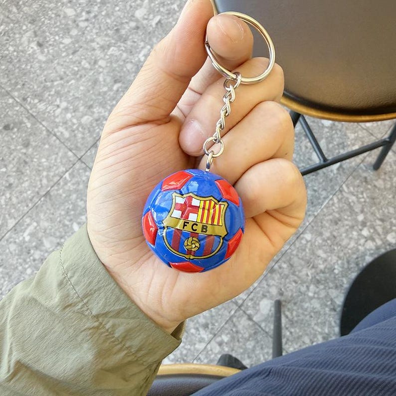 Puede incluir: Un llavero esf&eacute;rico con un patr&oacute;n de bal&oacute;n de f&uacute;tbol azul y rojo y el escudo del FCB impreso en la parte frontal. El emblema muestra una cruz roja, franjas verticales amarillas y rojas, y las letras FCB en negro sobre un fondo dorado.