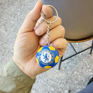 Puede incluir: Un llavero redondo con un dise&ntilde;o de bal&oacute;n de f&uacute;tbol en miniatura con paneles azules y amarillos. El centro del bal&oacute;n muestra el logotipo del Chelsea Football Club, que incluye un le&oacute;n azul sobre fondo blanco con el texto Chelsea Football Club.