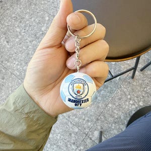 Puede incluir: Un llavero de metal con un peque&ntilde;o dije esf&eacute;rico de bal&oacute;n de f&uacute;tbol con paneles de color azul claro y blanco. El centro del bal&oacute;n muestra el logotipo del club de f&uacute;tbol Manchester City en azul, blanco y dorado, con el texto Manchester City.